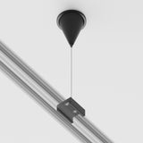 The 3-Phase Surface-Mounted Track Trinity Suspension Cable in black features a sleek cone-shaped ceiling mount with an adjustable cable, suspending a metal rail with two rectangular grooves, creating a modern contrast against the white ceiling as part of a track lighting system.