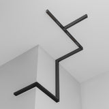 The Exility Magnetic Surface-Mounted Track 1m in black, featuring a linear minimalist design and customizable 48V setups, is installed on a white ceiling and wall, creating a zigzag pattern across the rooms corner.