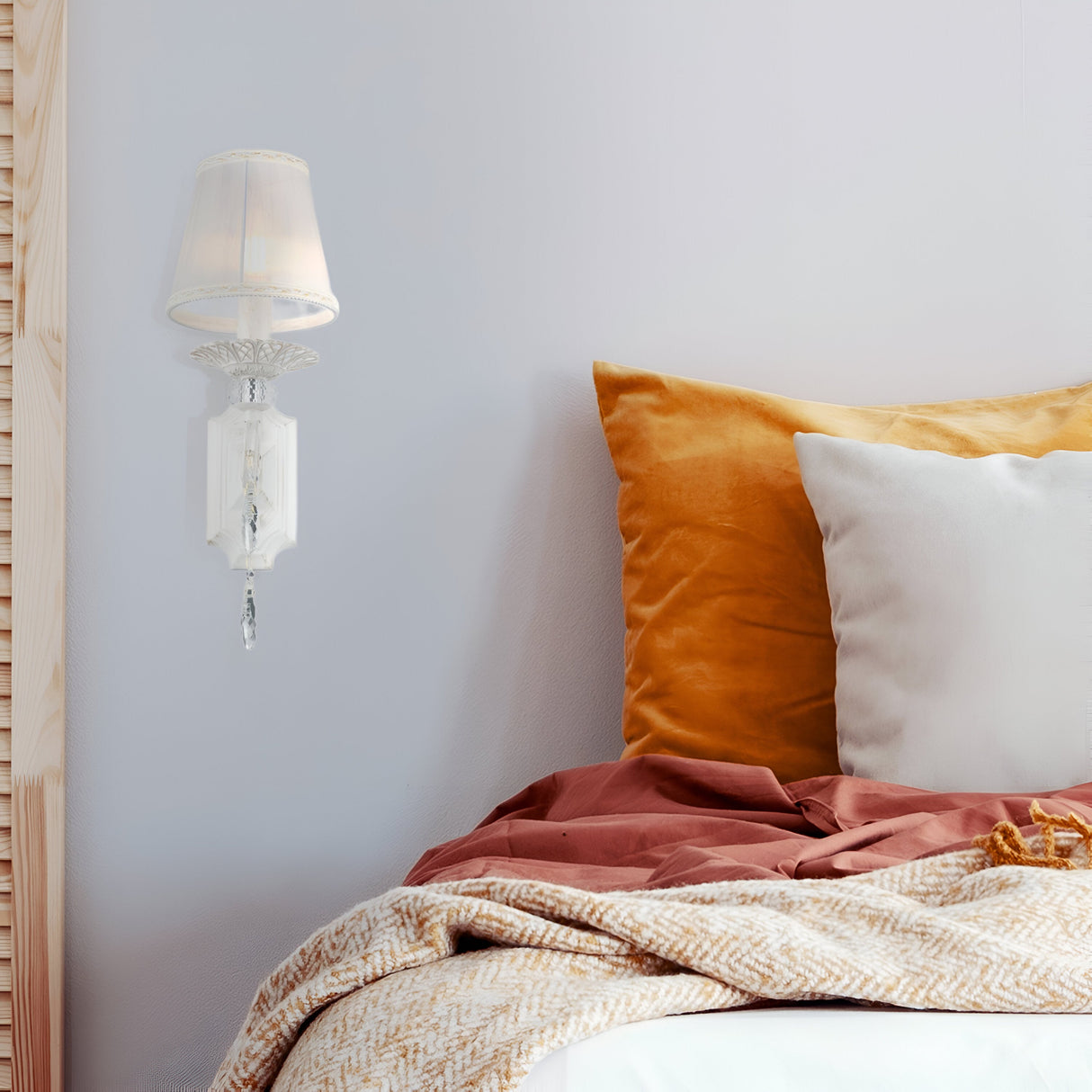 A cozy bedroom features the Grace Wall Light - White & Gold With Fabric Shade, complemented by mustard, brown, and white pillows on the bed, which is covered with a white and beige patterned blanket for a warm ambiance.