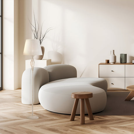 The minimalist living room features a round white sofa, a small wooden stool, and the Grace Floor Lamp - White with a pleated shade. Decorative vases on a low wooden cabinet complement the cream-colored wall with modern art, all bathed in light from tall windows.