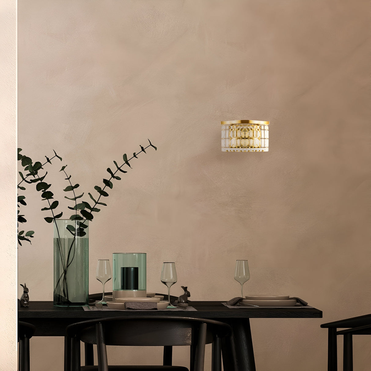 A minimalist dining scene showcases a dark table paired with black chairs, adorned with wine glasses and a vase of tall eucalyptus branches. The Luxe Classic style is enhanced by the Dune 2 Light Crystal Wall Light - Gold, casting light on the neutral textured wall behind.