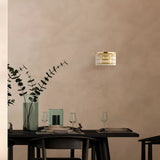 A minimalist dining scene showcases a dark table paired with black chairs, adorned with wine glasses and a vase of tall eucalyptus branches. The Luxe Classic style is enhanced by the Dune 2 Light Crystal Wall Light - Gold, casting light on the neutral textured wall behind.