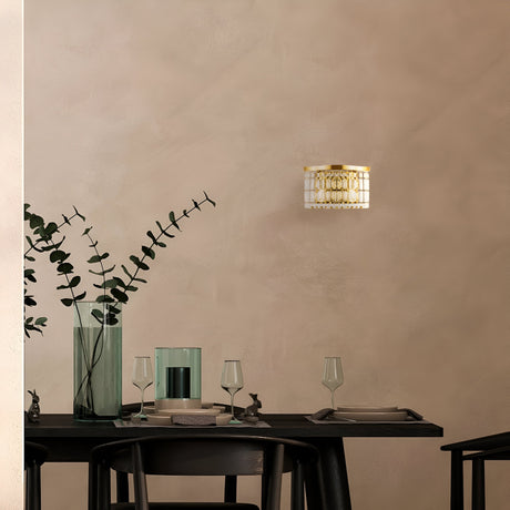 A minimalist dining scene showcases a dark table paired with black chairs, adorned with wine glasses and a vase of tall eucalyptus branches. The Luxe Classic style is enhanced by the Dune 2 Light Crystal Wall Light - Gold, casting light on the neutral textured wall behind.