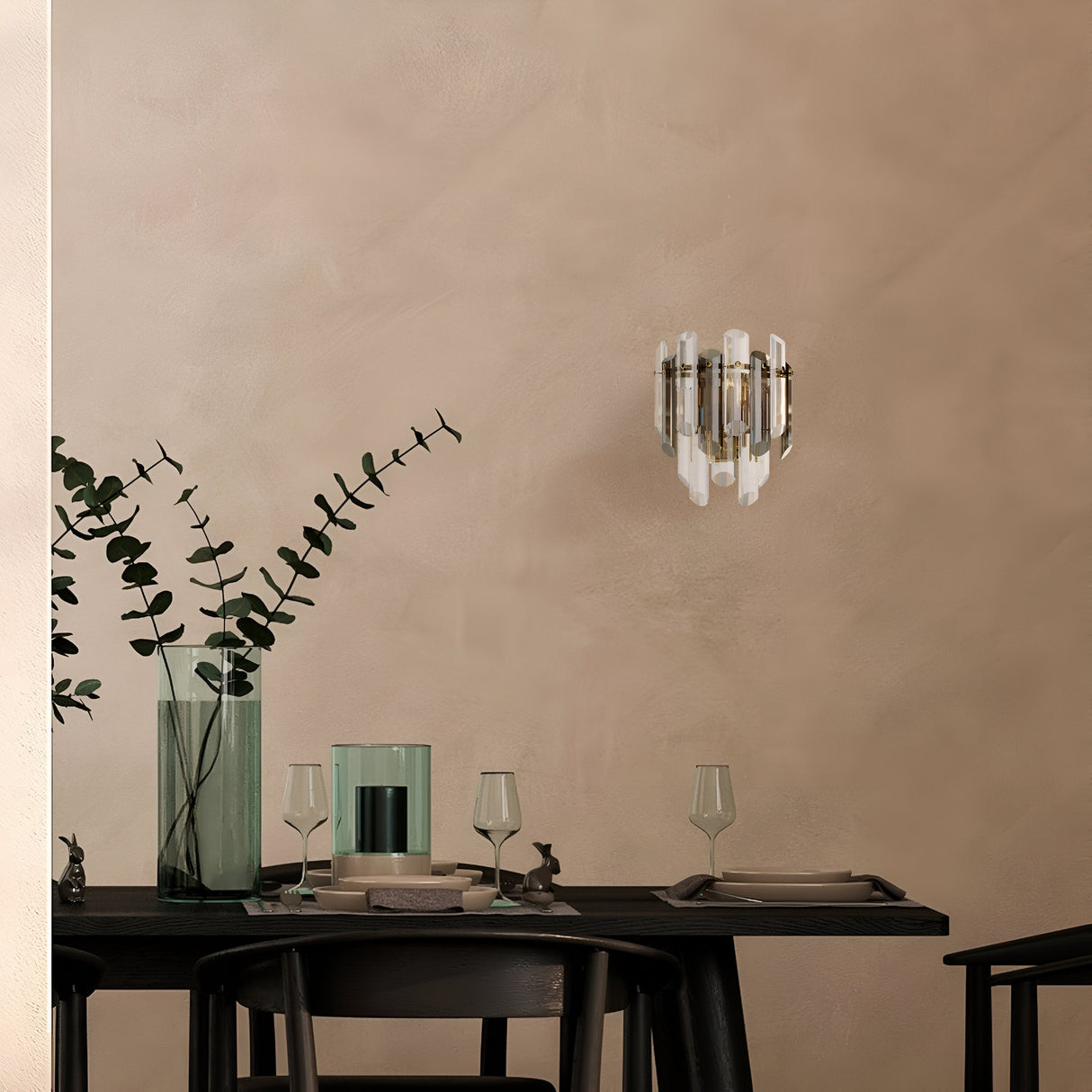 A minimal dining area boasts a dark wooden table for two, adorned with glassware and a vase holding eucalyptus branches. The beige walls are softly illuminated by the Flare 2 Light Wall Light - Gold With Crystal. Simple black chairs complement the setup.