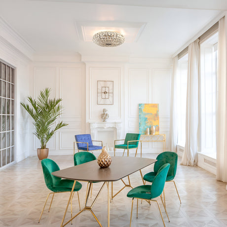 The elegant dining room features a modern table accompanied by teal and blue chairs. The Broche 6 Light Crystal Chandelier - Nickel adds luxurious lighting. A potted plant, artwork, and centerpiece beautify the space, while large windows with sheer curtains allow natural light to illuminate the room.