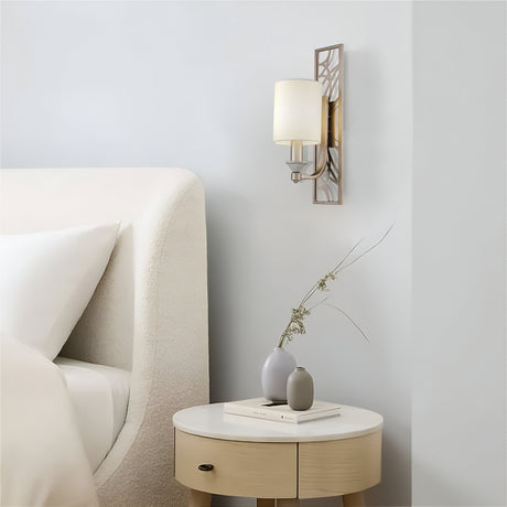 A modern bedroom showcases a white curved headboard and a small round bedside table with a gray vase of dried flowers and stacked books. The Vittoria Wall Light - Gold With Cream Fabric Shade softly casts ambient lighting on the neutral-toned wall.