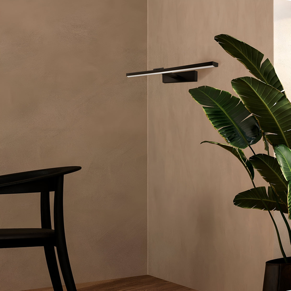 In a minimalist interior, a black chair sits beside the Bris LED Picture Light 12W 4000K - Black & White for modern energy-efficient illumination. A tall green plant with broad leaves complements the beige walls and wooden flooring.