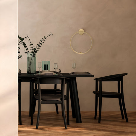 A modern dining area features a black table set for two with greenery and candles, surrounded by two black chairs. The Halo LED Medium Ring Wall Light 36W 4000K in brass adds a minimalist touch on the wall, while wood flooring complements the decor.