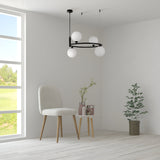 In a minimalist room with light wood flooring, a round white side table holds vases and a plant. Nearby, a white chair with wooden legs complements the decor. The focal point is the Ring 4 Light Circular Suspension in black with white glass, while large windows showcase the green landscape outside.