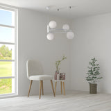 A minimalist room features a cushioned chair beside a small round table with books and vases. A Ring 4 Light Circular Suspension - White With White Glass hangs above. To the right, a potted plant rests on the wooden floor, and a large window offers views of green fields.