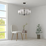 A minimalist room features a cushioned chair and a round table with potted plants. The Alicante 6 Light Chandelier - Nickel adds elegance as sunlight floods through the large window, framing lush greenery and epitomizing modern interiors.