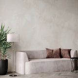 A minimalist living room includes a beige sofa with two brown cushions, an Alicante Floor Lamp - Nickel, and a potted green plant by the textured beige wall. A small round side table is positioned to the right of the sofa.