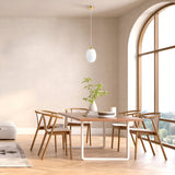 A minimalist dining room showcases a light wooden table with six matching chairs on a herringbone floor. Above, an Aura Pendant Light - Brass With Inner Frosted & Outer Clear Glass Shade illuminates the space, highlighting a potted plant on the table. A large arched window offers an inviting view outside.