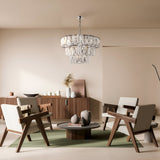 A modern living room showcases four wooden chairs around a circular coffee table. Above, the Puntes 18 Light Chandelier in chrome adds elegance. The room includes a sideboard with decor on a light floor and a green oval rug, all set against neutral walls.