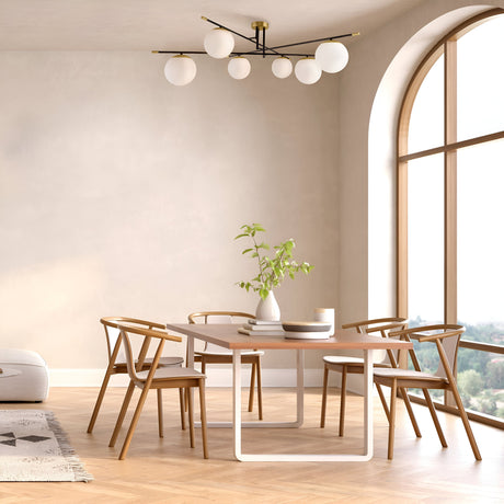 A modern dining room showcases a wooden table with six chairs, accented by a white vase with green branches. Natural light floods through a large arched window, complemented by the Nostalgia 6 Light Semi-Flush fixture in matte black and gold for a contemporary touch above.
