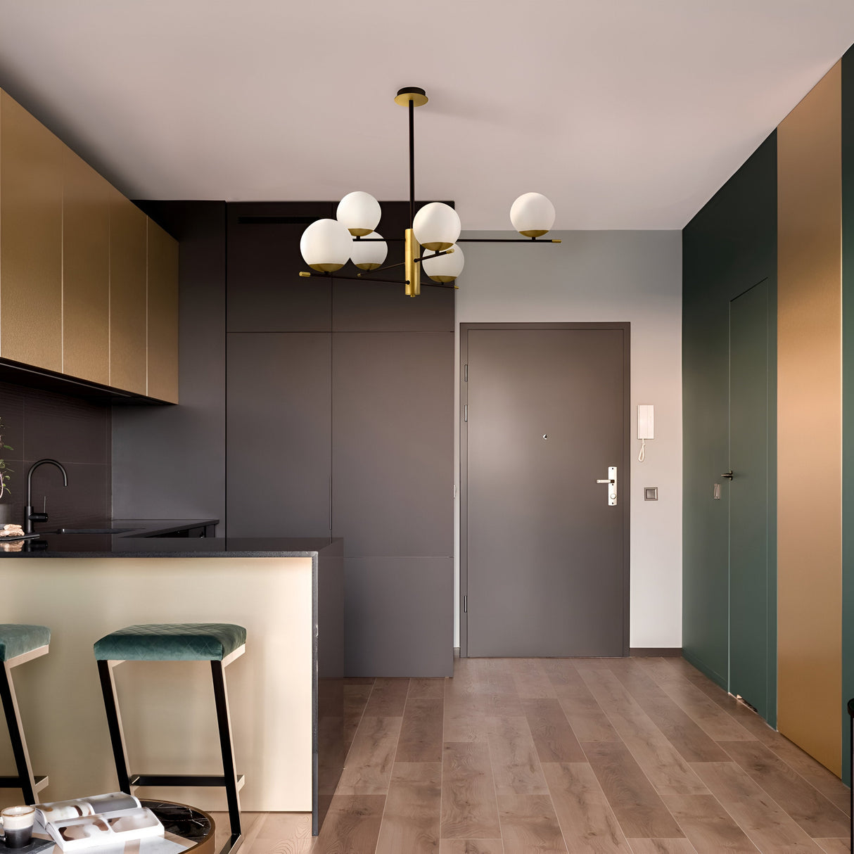 A modern minimalist kitchen features a gray door, gold cabinetry, and a dark counter. A Nostalgia 6 Light Chandelier - Gold hangs over wooden flooring. Two bar stools with teal cushions are positioned by the light-colored kitchen island.