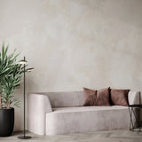 A minimalist living room features a light gray sofa with two brown cushions, a tall potted plant on the left, and the energy-efficient Fad LED Floor Lamp - Black. A small round table with a wireframe base sits to the right, all set against a textured neutral wall.