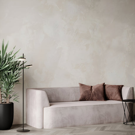 A minimalist living room features a light gray sofa with two brown cushions, a tall potted plant on the left, and the energy-efficient Fad LED Floor Lamp - Black. A small round table with a wireframe base sits to the right, all set against a textured neutral wall.