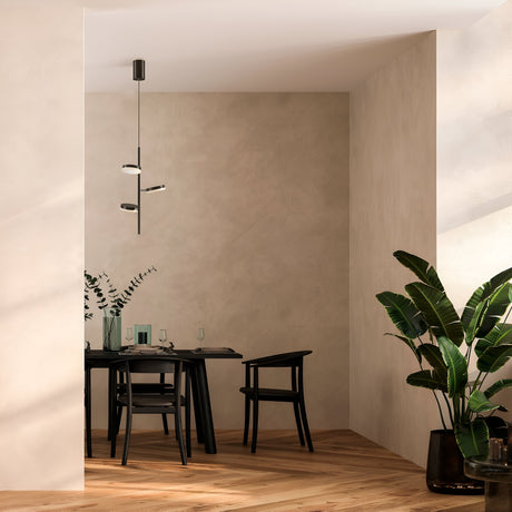 A minimalist dining area features a small black table for two with elegant black chairs beneath the Fad Adjustable LED Pendant in black. A vase of foliage adorns the table, large potted plants sit in the corner, and warm wooden flooring completes the look.
