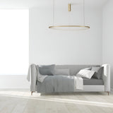 A minimalist living room features a light gray sofa with gray and white cushions and a draped throw against a white wall. The Glint LED Circular Suspension Light 80cm - Brass provides energy-efficient lighting, while the serene space is completed with light wood flooring.