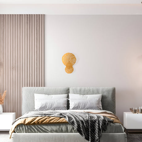 A modern bedroom displays a grey upholstered bed with checkered pillows and a grey blanket. A textured beige wall features a matte brass decorative piece, while a small white nightstand holds a plant and candle, warmly lit by the Lovetann Wall Light - Gold With Perforated Metal Discs.