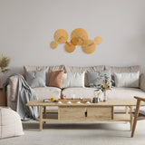 A cozy living room showcases a light beige sofa with gray and orange patterned cushions. A wooden coffee table displays tea cups and decor, while abstract orange wall art above the sofa is illuminated by a Lovetann 4 Light Wall Light in gold. Dried plants grace the corner.