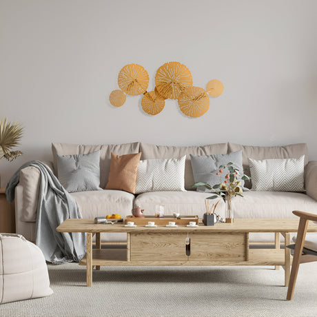 A cozy living room showcases a light beige sofa with gray and orange patterned cushions. A wooden coffee table displays tea cups and decor, while abstract orange wall art above the sofa is illuminated by a Lovetann 4 Light Wall Light in gold. Dried plants grace the corner.