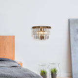 A modern bedroom features a wooden bed with gray bedding and the Revero Classic Wall Light in brass with transparent glass on the white wall. Two glass pots with green plants are on the floor, while a large framed artwork leans casually against the wall.