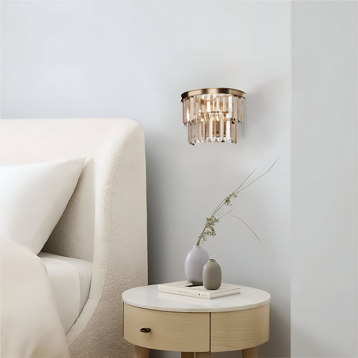 A minimalist bedroom features a white bed with a plush headboard, a round wooden nightstand adorned with a small vase and books, and the Revero Classic Wall Light in gold with amber glass that emits soft light against the light gray wall.