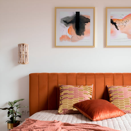 The bedroom features an orange upholstered headboard with two patterned pillows and an orange pillow. Above, abstract framed artworks are flanked by Facet Wall Lights - Gold. A small plant rests on the nightstand nearby.