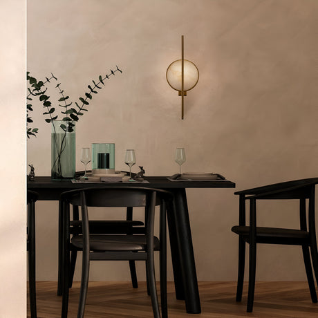 A minimalist dining area showcases a dark wooden table for two, adorned with glassware and cutlery. A tall vase with greenery adds freshness, while the Marmo Wall Light - Gold & Natural Stone subtly illuminates the beige wall. Two black chairs complete this modern interior design setting.