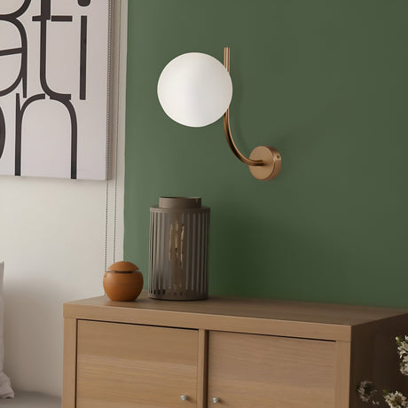 A modern interior showcases a wooden cabinet with a small brown vase and tall gray candle holder. The dark green wall is illuminated by the Rendez-vous Wall Light - Brass With White Opal Glass, as a large abstract art piece subtly comes into view.