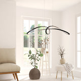 A minimalist room with large windows, light walls, and a Curve LED Loop Suspension Light - Black as the centerpiece. It includes a cushiony chair, potted plants, and a wooden stool adorned with a vase and decorative branches.