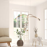 A minimalist, sunlit room boasts the Curve LED Loop Suspension Light in gold with an integrated LED. White walls and large windows enhance the spaciousness, complemented by a beige chair, two potted plants, and a small table with branches on the light wood floor.