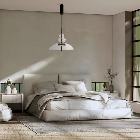 A minimalist bedroom features a large bed with white bedding and pillows, flanked by two decorative nightstands. The Bangkok LED Pendant Light 40cm in black with a transparent glass shade hangs above. Sunlight pours through a large right-side window, enhancing the modern design.