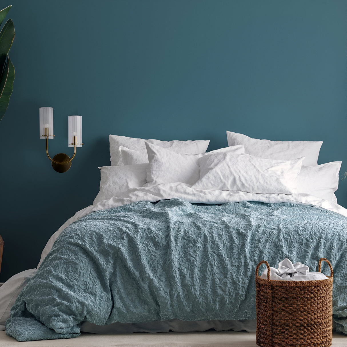 A cozy bedroom features a blue accent wall and a neatly made bed with white pillows and a textured blue blanket. A woven basket of towels sits on the floor, while the Arco 2 Light Wall Light - Brass With Ribbed Glass adds charm on the left. A plant enhances the mid-century modern feel.