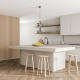 A minimalist kitchen features a large white island with three wooden stools and a fruit bowl with lemons. Gray cabinets and light wood open shelves adorn the back wall. The Palette Multicoloured Pendant Light - Grey adds flair, while a herringbone wood pattern enhances the floor.