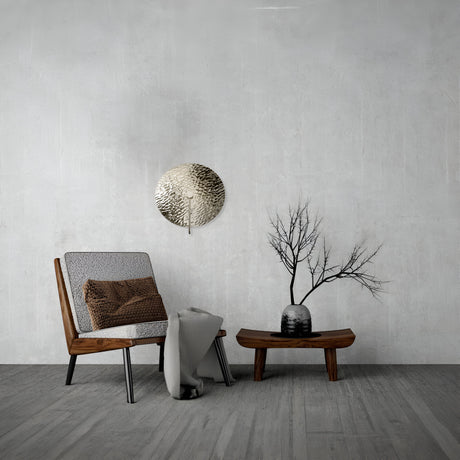 A modern design graces this minimalist living room, featuring a wooden chair with a cushion and blanket, a low wooden table, and a vase of dried branches. The Mare Wall Light with chrome finish and round mirrored glass diffuser highlights the metal disc on the gray wall, complemented by the light hardwood floor.