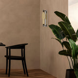 A modern interior includes a black chair and a large potted plant with broad green leaves in the corner, while the Verticale LED Wall Light 3000K 8W - Chrome with Cognac Ribbed Glass emits a soft glow. Light, warm-toned walls and floors create an inviting atmosphere.