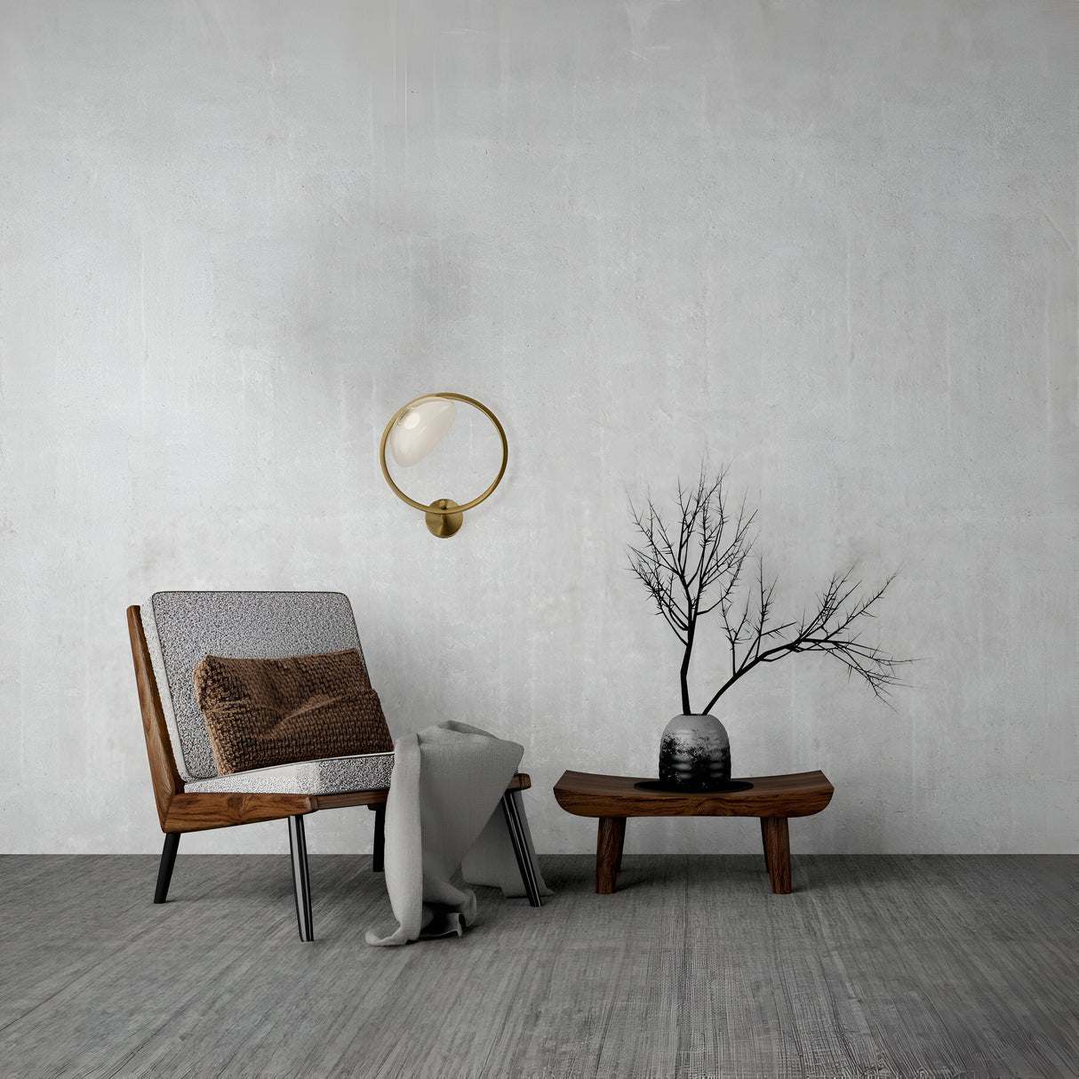 A minimalist room features a modern wooden chair with a brown cushion and draped gray blanket. A small wooden table supports a black-and-white vase with bare branches. The Lunare LED Downward Wall Light in brass illuminates the plain gray wall above textured flooring, epitomizing modern elegance.