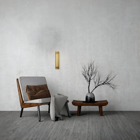 A minimalist living room features a wooden chair with a brown cushion, gray throw blanket, and wooden coffee table adorned with a vase of bare branches. The wall boasts the energy-efficient Sonata LED Wall Light 12W 3000K in brass against a gray textured surface.