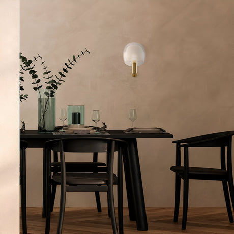 A minimalist dining area showcases a dark wood table set for two, featuring clear glasses and a tall vase of eucalyptus. Surrounded by black chairs, the Vision Wall Light - Gold with Inner Frosted & Outer Clear Glass adds elegance to the beige wall, enhancing the rooms subtle charm.