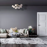 A living room features a modern gray sofa, assorted pillows, and a round coffee table with vases of dried flowers. A Dallas 12 Light Semi-Flush fixture in brass with white glass hangs elegantly above. Gray walls, a textured rug on dark wood floors, and a white-paneled door complete the space.