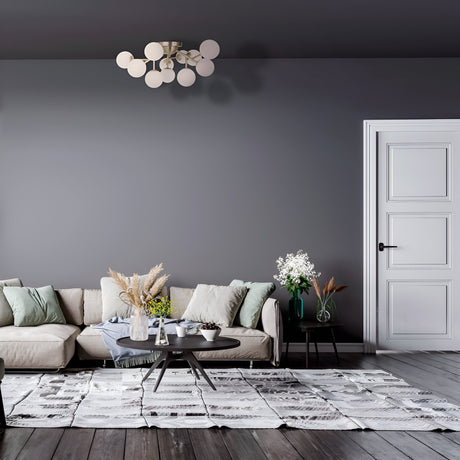A living room features a modern gray sofa, assorted pillows, and a round coffee table with vases of dried flowers. A Dallas 12 Light Semi-Flush fixture in brass with white glass hangs elegantly above. Gray walls, a textured rug on dark wood floors, and a white-paneled door complete the space.