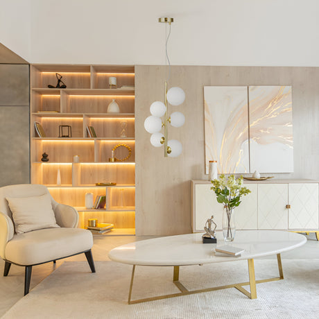 A modern living room features a beige armchair, an oval white coffee table with a flower vase, and a sideboard. The Dallas 6 Light Drop Suspension in gold with white glass spheres hangs above. Built-in shelves on the wall feature ambient lighting and abstract art in gold frames.