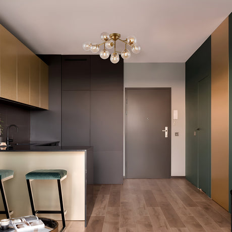 The modern kitchen and entryway boast sleek dark gray and gold cabinetry, highlighted by the Dallas 12 Light Semi-Flush - Gold With Amber Glass chandelier. Wooden flooring supports two bar stools at the counter, and a gray entry door with intercom is accented by gold metal fittings.