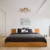 A minimalist bedroom features a wooden bed with dark gray bedding and neutral pillows. The Dallas 20 Light Semi-Flush in gold with amber glass hangs from the ceiling, while a modern abstract painting adorns the white wall. Light wooden flooring complements the decor nicely.