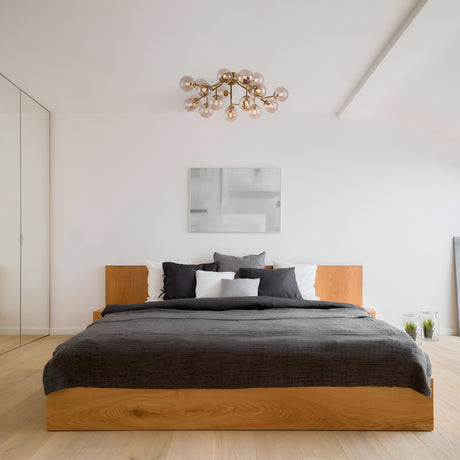 A minimalist bedroom features a wooden bed with dark gray bedding and neutral pillows. The Dallas 20 Light Semi-Flush in gold with amber glass hangs from the ceiling, while a modern abstract painting adorns the white wall. Light wooden flooring complements the decor nicely.