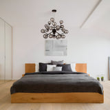 Modern bedroom featuring a wooden bed frame, gray bedding, and multiple pillows. A Dallas 24 Light Chandelier in Black with Smoky Glass adds ambiance above. Minimalistic art adorns the white wall, complementing the light wooden floor and glass closet doors on the left.