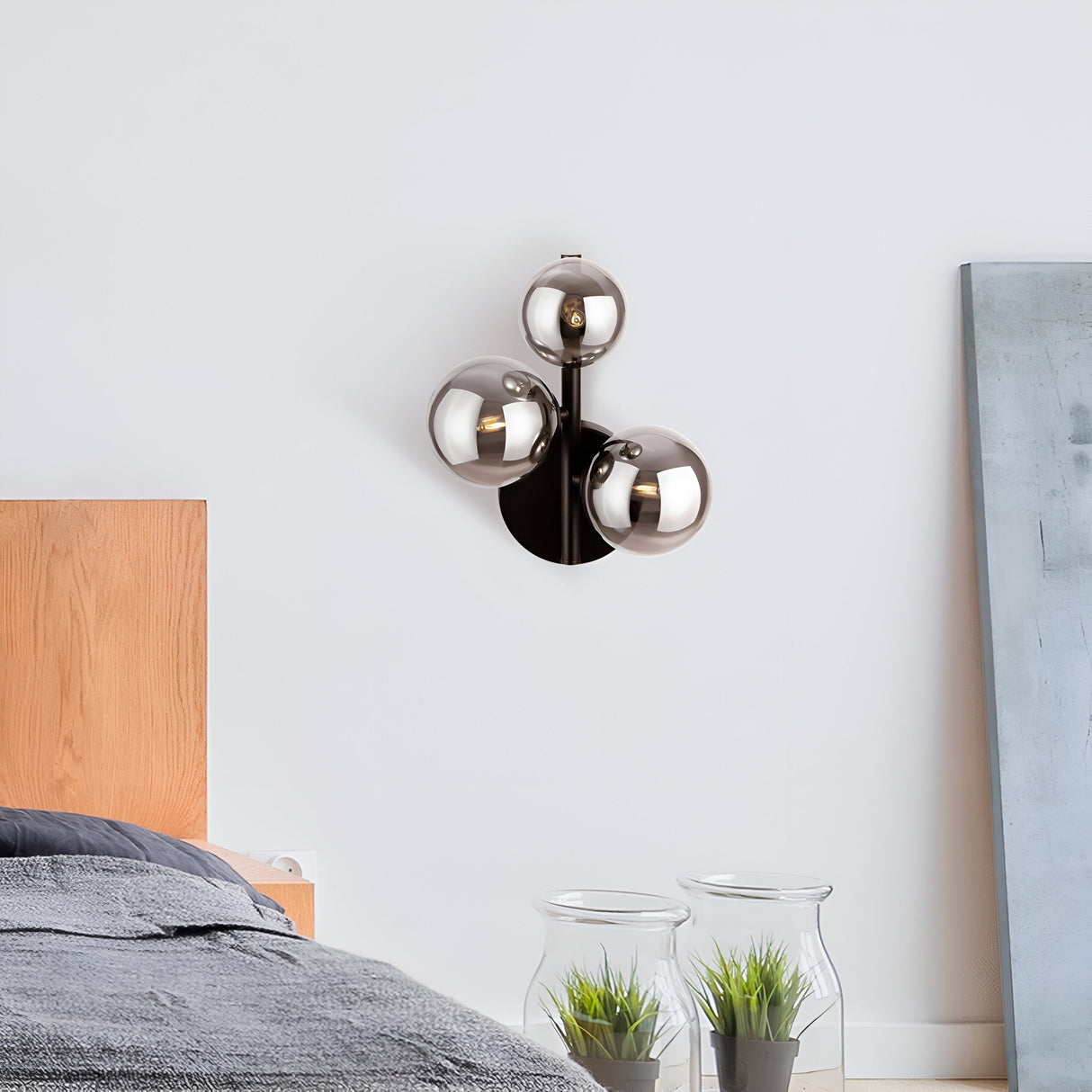 A modern bedroom showcases a wooden bedframe with gray bedding. The Dallas 3 Light Wall Light - Black With Smoked Mirrored Glass adds elegance to the white wall, complemented by two glass vases with green plants and a large canvas leaning stylishly for contemporary charm.